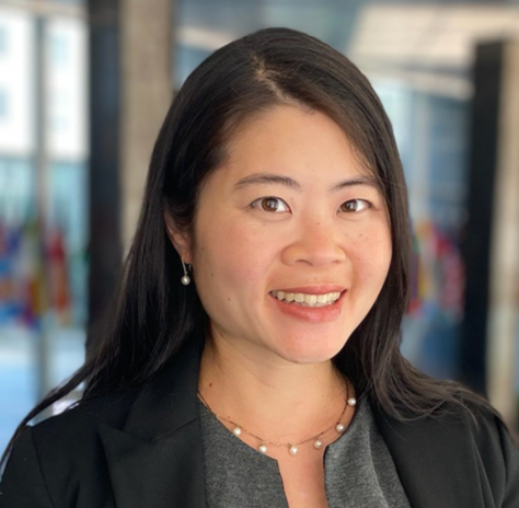 Arisa is Asian with a peachy undertone and has black straight long hair. She wears a dark grey dress with a black blazer and a pearl necklace and earrings, standing in front of the flags in the State Department lobby.