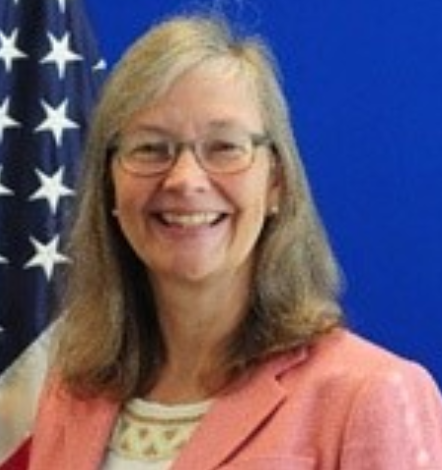Ms. Kay, a white woman, smiles in front of a U.S. flag and blue background. She is middle-aged with shoulder-length straight auburn hair and wearing blue glasses, pearl earrings, and a salmon blazer over a beaded white blouse.