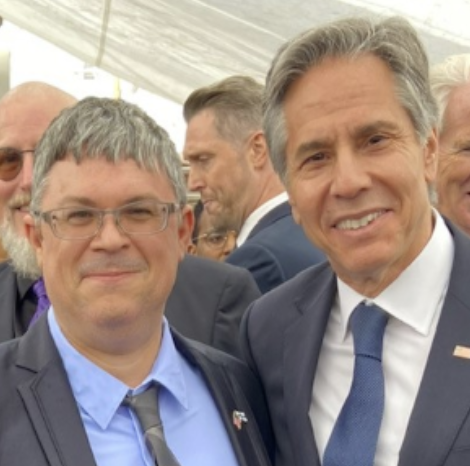 Doug poses with Secretary Blinken: both gentlemen stand shoulder-to-shoulder, grin at the camera, and wear dark jackets, dress shirts, ties, and flag pins. Doug is white with brown eyes, silvery hair, and glasses.