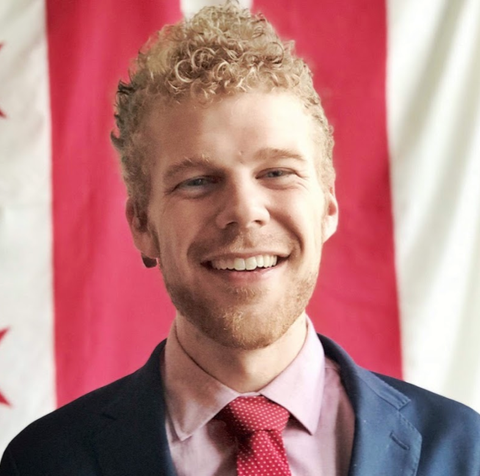 Robb is white with curly blond hair, light orange facial hair, a pink shirt, and a red tie. A cochlear implant is visible on his right ear. He stands in front of the flag of Washington, DC.