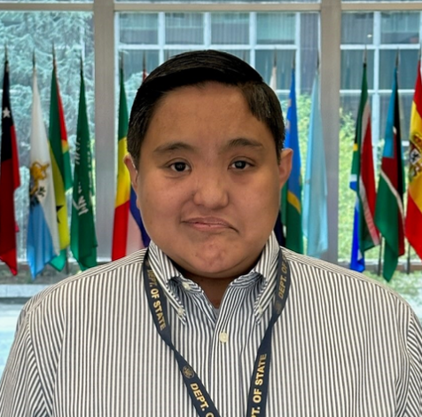 Wayne has brown skin and brown eyes, straight black hair, a white and gray striped dress shirt, and a State Department lanyard. They pose in front of the flags in the State Department lobby.