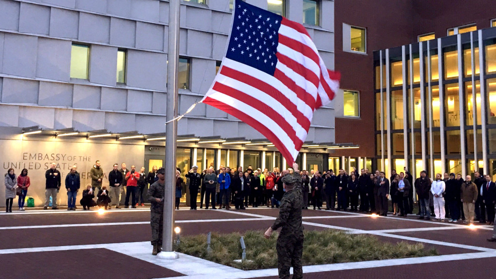 An image of a United States flag being raised on a flagpole in front of a U.S Embassy. Below the flag, two soldiers in uniform are involved in the flag-raising ceremony. A crowd of people stands gathered behind them, observing the event.